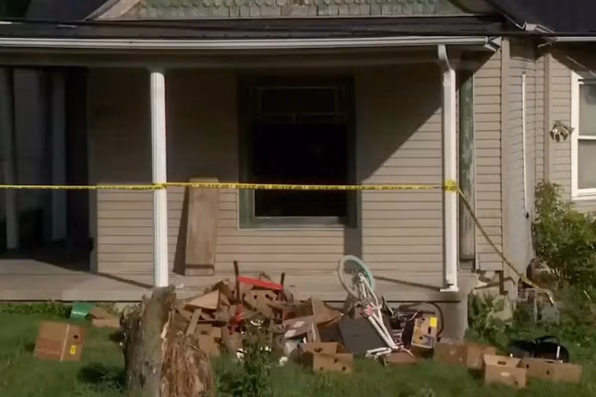 The image depicts a residential property cordoned off with yellow police tape. The house, characterized by a sloped roof and a front porch supported by white pillars, appears to be the scene of an incident. Several items, including debris and garden elements, are scattered around the front yard, suggesting some form of disturbance or investigation. The windows are darkened, and the front door is closed.