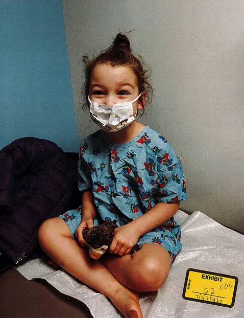 A young child in a hospital gown and mask, sitting cross-legged and holding a stuffed animal.