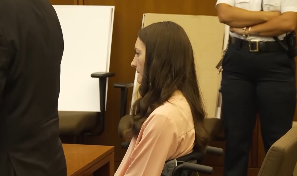 A woman with long brown hair, wearing a pink shirt, sits in a chair in a courtroom.