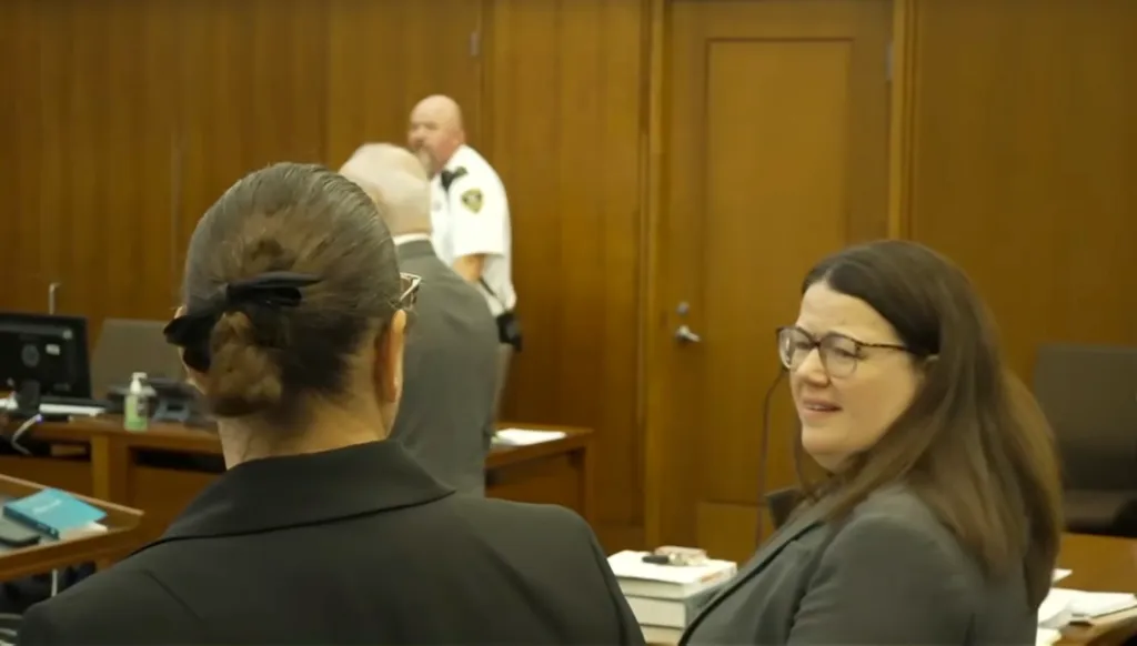 Two women in a courtroom, one with her back to the camera, and the other wearing glasses and looking distressed.