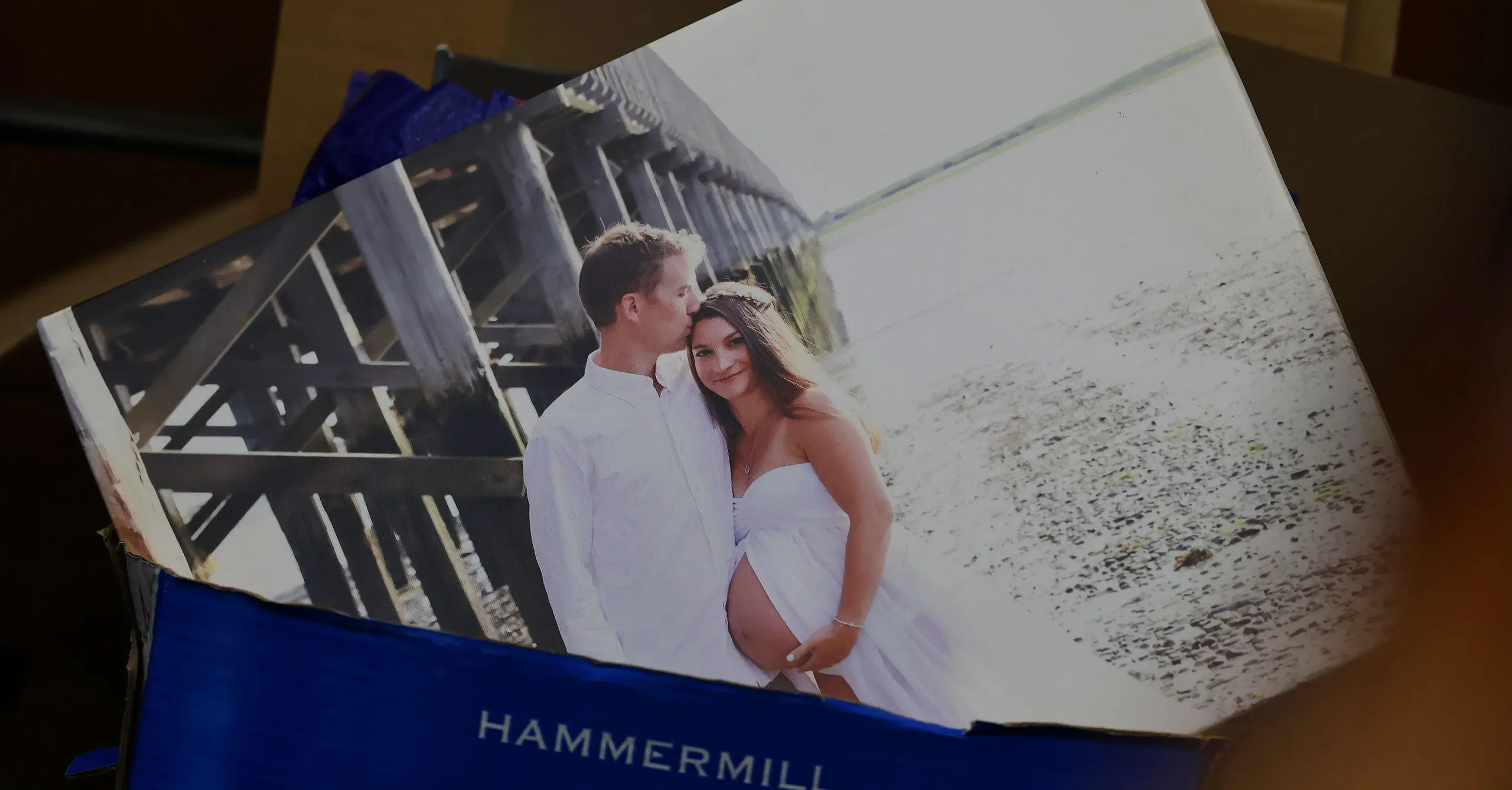 Printed maternity photo of a couple in white clothing near a wooden pier on a beach, resting on a Hammermill paper box.