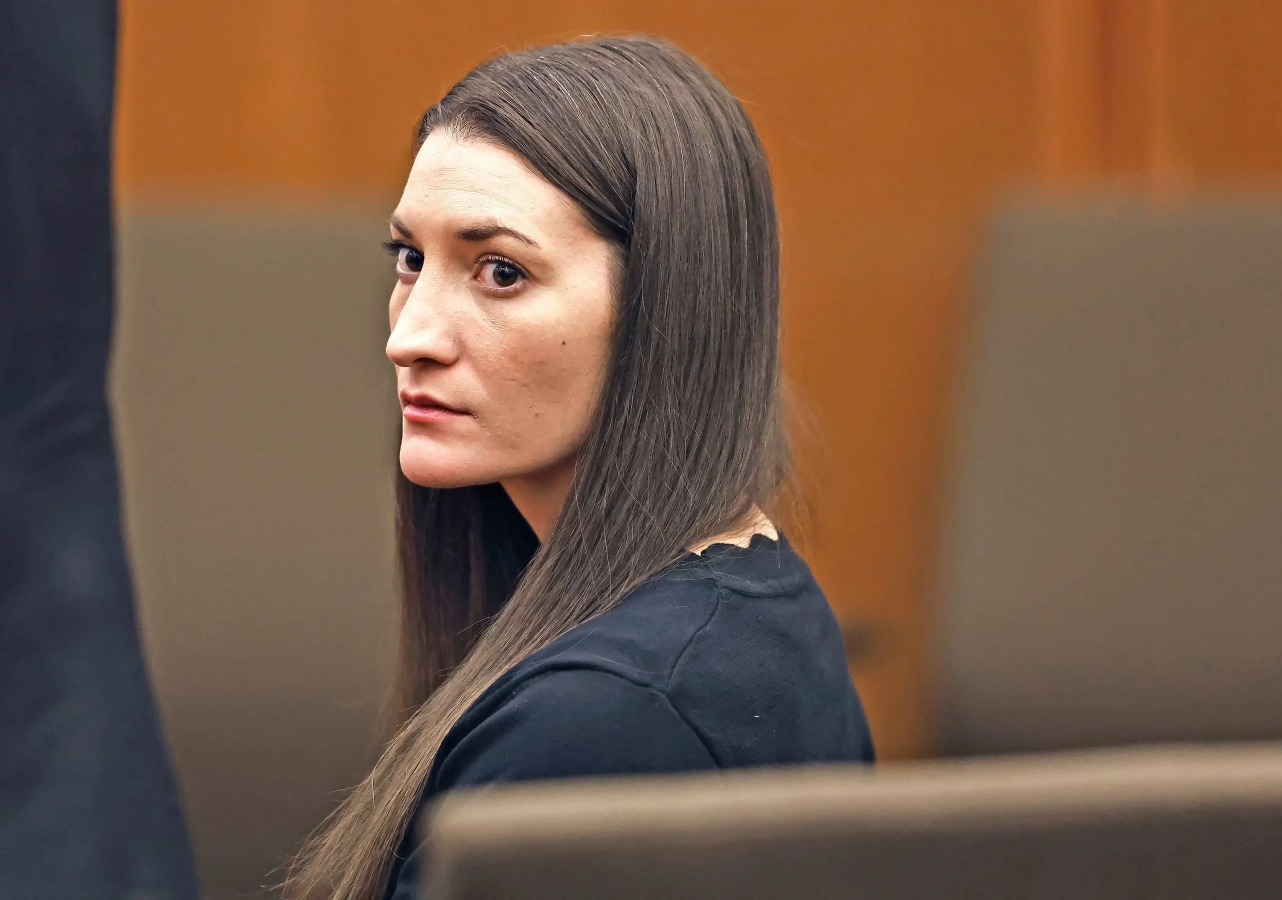 Woman with long dark hair in a black top seated in a courtroom, looking sideways toward the camera.