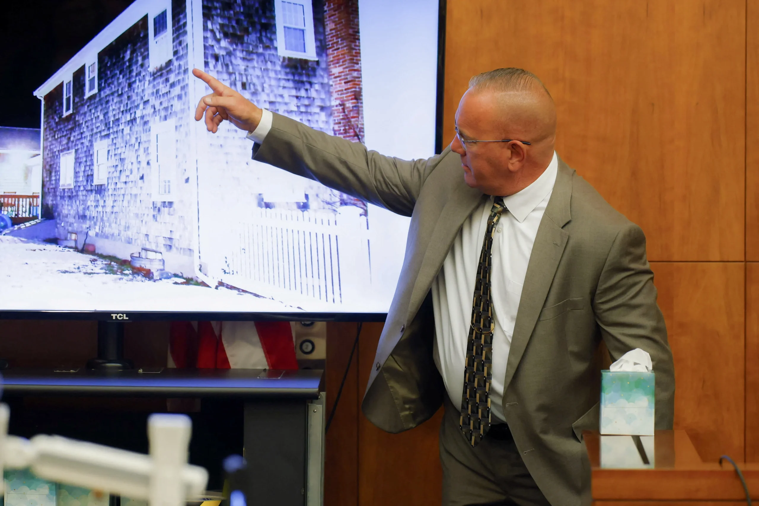 A man in a gray suit points at a large screen showing a photo of a house exterior while testifying in a courtroom.