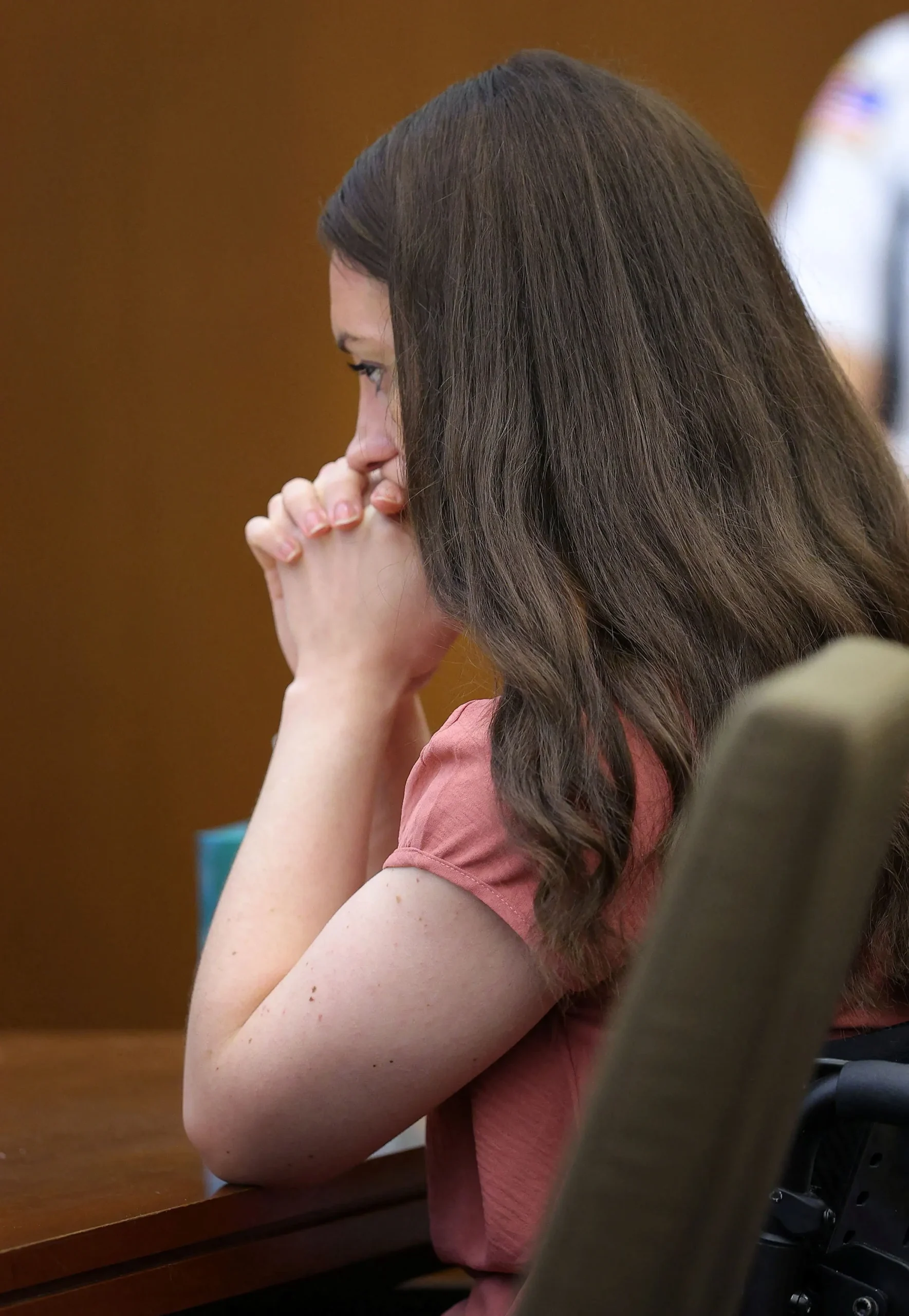 Woman with long brown hair in a pink top seated at a courtroom table, hands clasped over her mouth, eyes downcast.