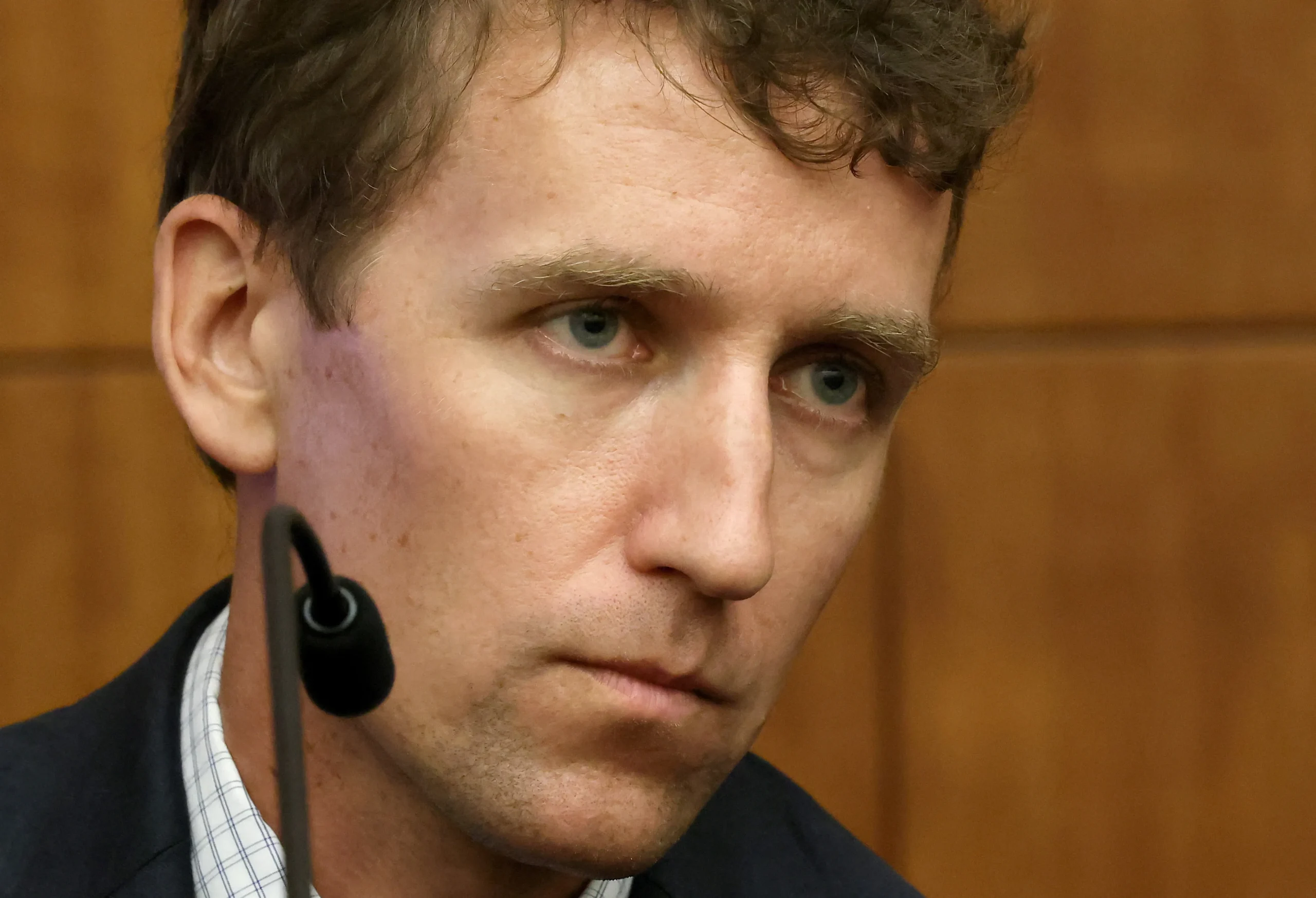 Close-up of a man with curly brown hair and blue eyes wearing a lapel microphone, seated in a courtroom witness stand.