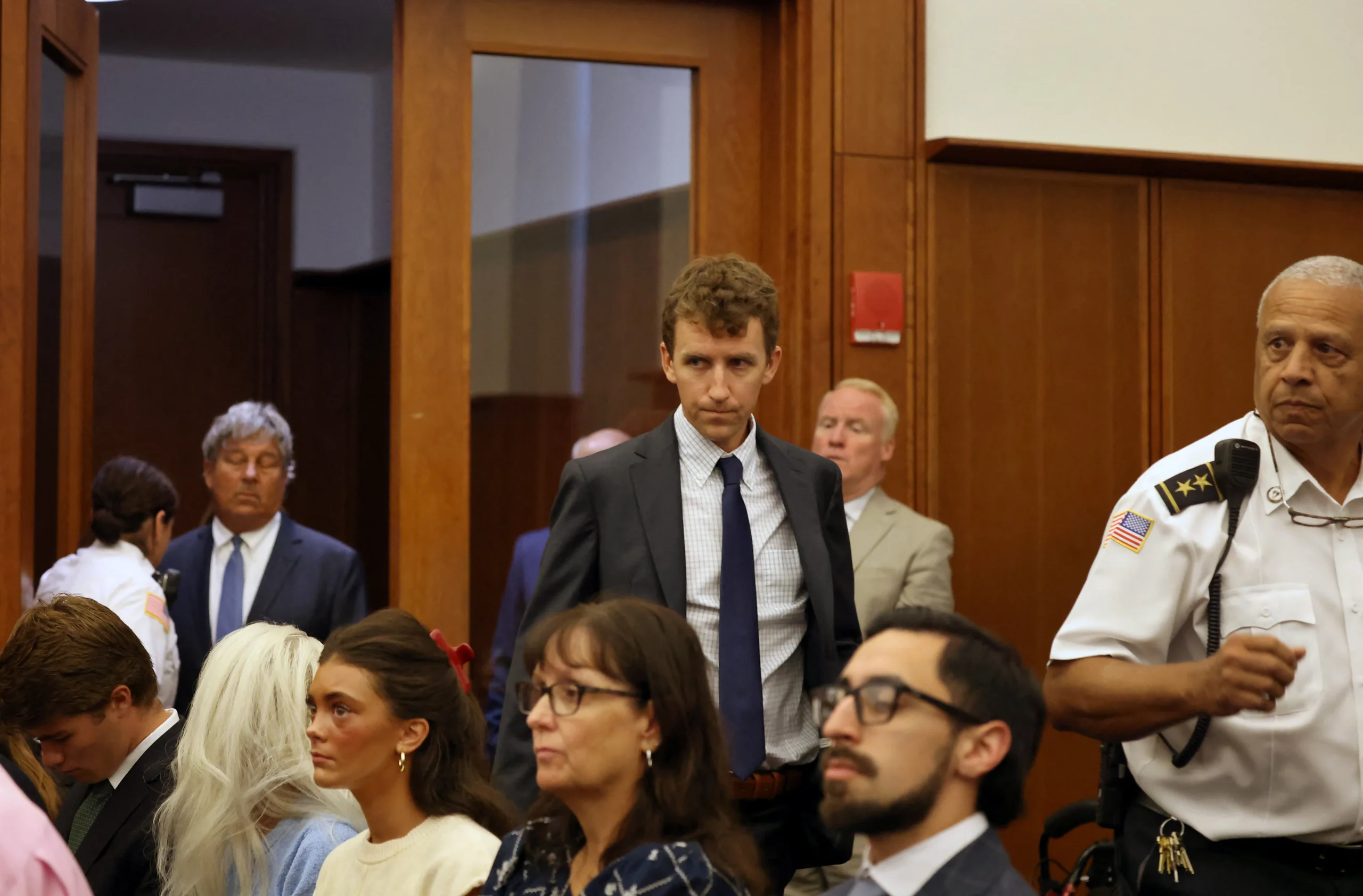 A man in a gray suit and blue tie walks through a crowded courtroom toward his seat, surrounded by other attendees and a court officer.