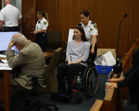 A woman in a wheelchair is escorted to a table in a courtroom