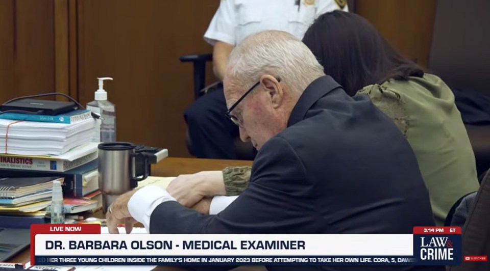 A balding man in glasses with a green-clad woman seated behind him in a courtroom.