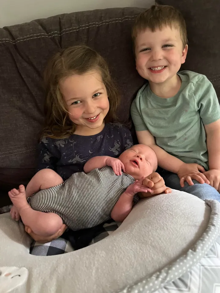 Three young children, including a newborn, sitting together on a couch, smiling.