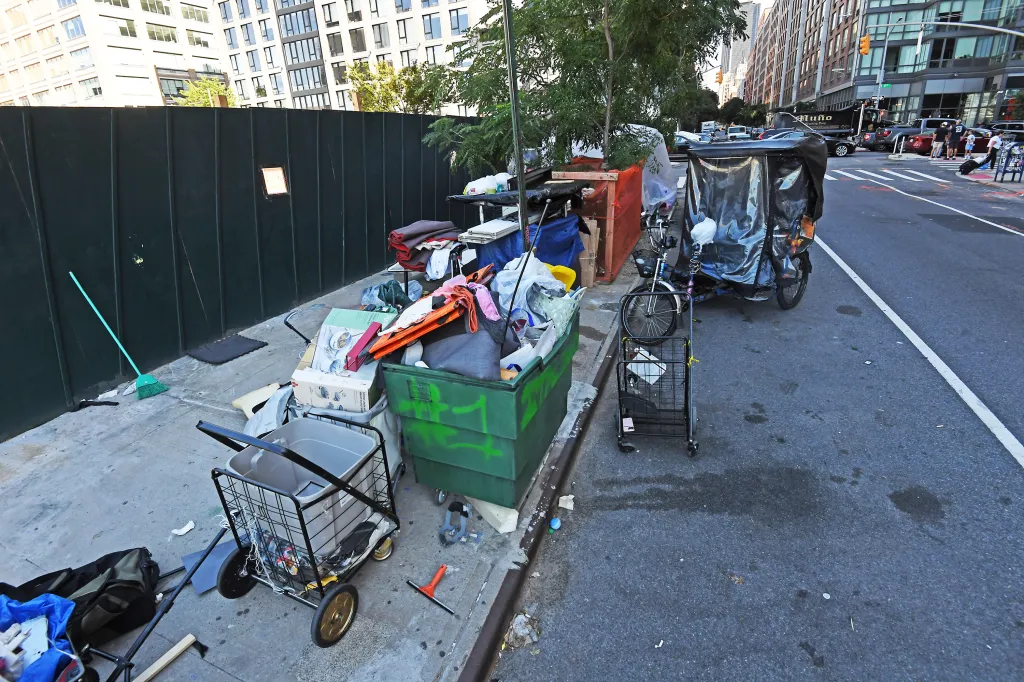 An urban encampment with belongings piled on the sidewalk beside a busy street.