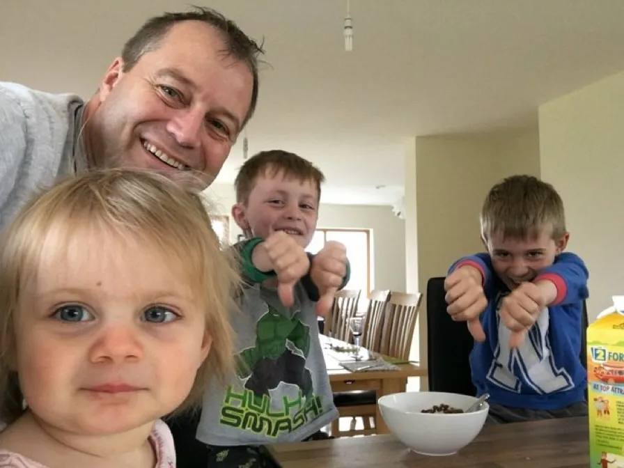 Andrew McGinley smiling with his three children, two boys giving thumbs down and a little girl looking at the camera.